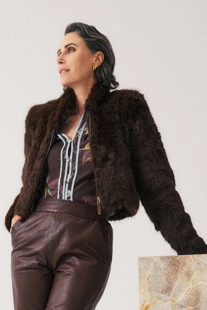 Woman wearing a brown fur jacket and leather pants against a plain background