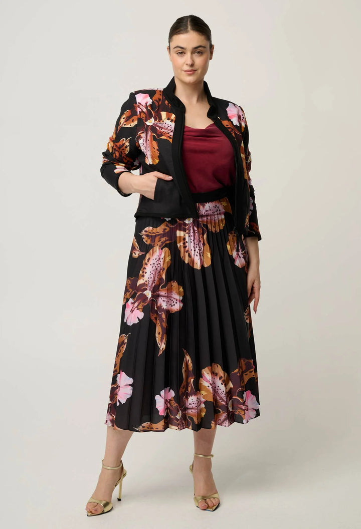 A woman modeling a black midi skirt and matching jacket set with a pink, brown, and orange floral print, worn over a burgundy top and paired with beige high-heeled sandals, against a light gray studio background.