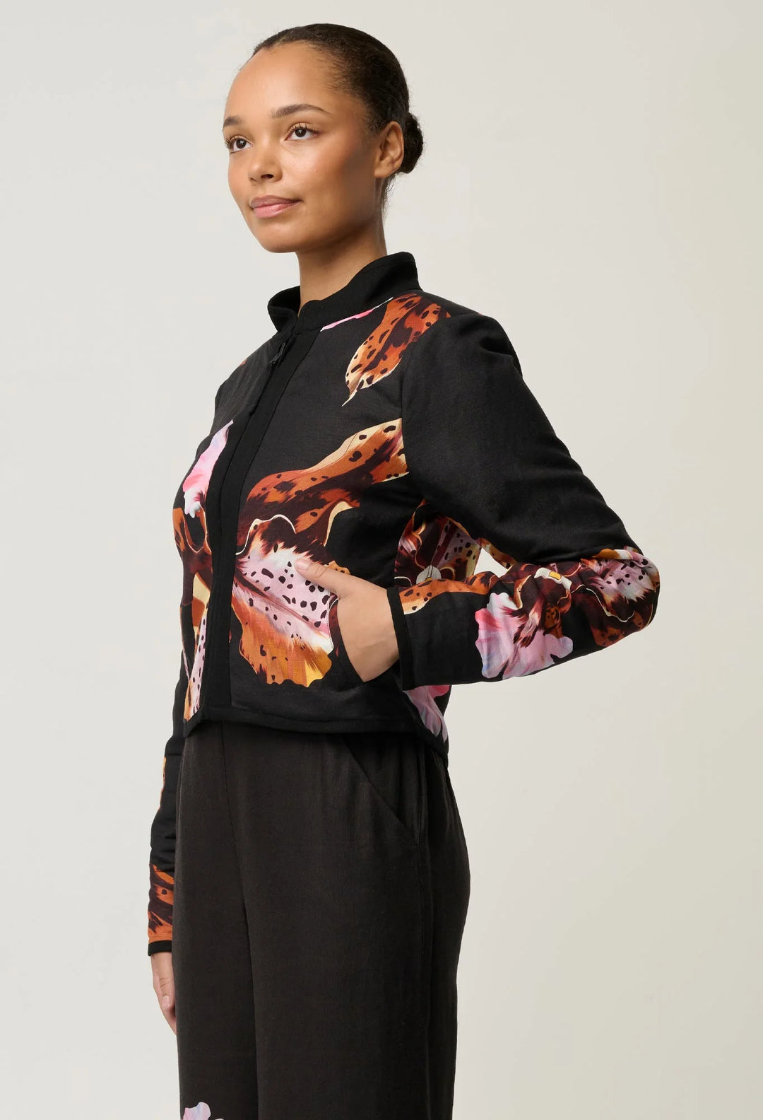 A woman modeling a black long-sleeve jacket with a vibrant abstract floral print in orange, pink, and brown, paired with black trousers, against a light gray studio background.