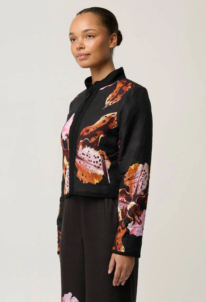 A model wearing a black cropped jacket with a bold orange, pink, and white abstract floral print, paired with black trousers, against a light gray studio background.