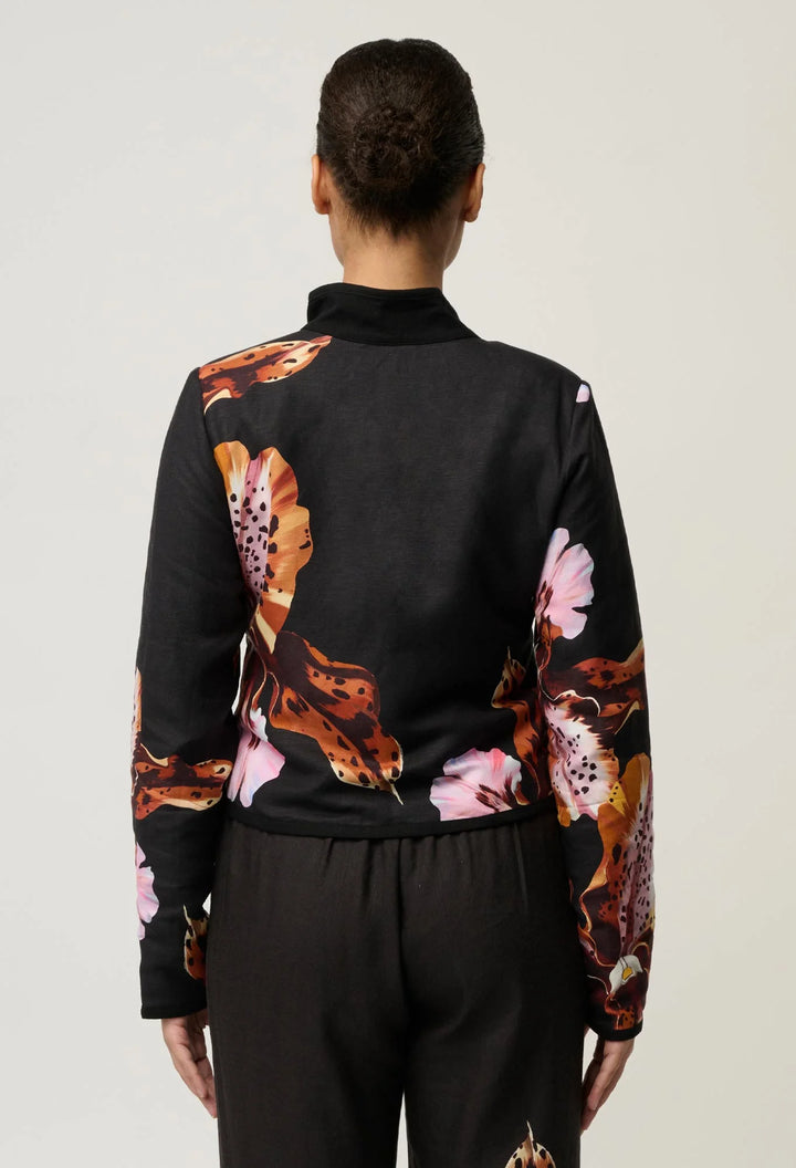 A model viewed from the back wearing a black long-sleeve mock-neck jacket with a bold orange, brown, and pink floral print on the sleeves and back, paired with black trousers, against a light gray studio background.