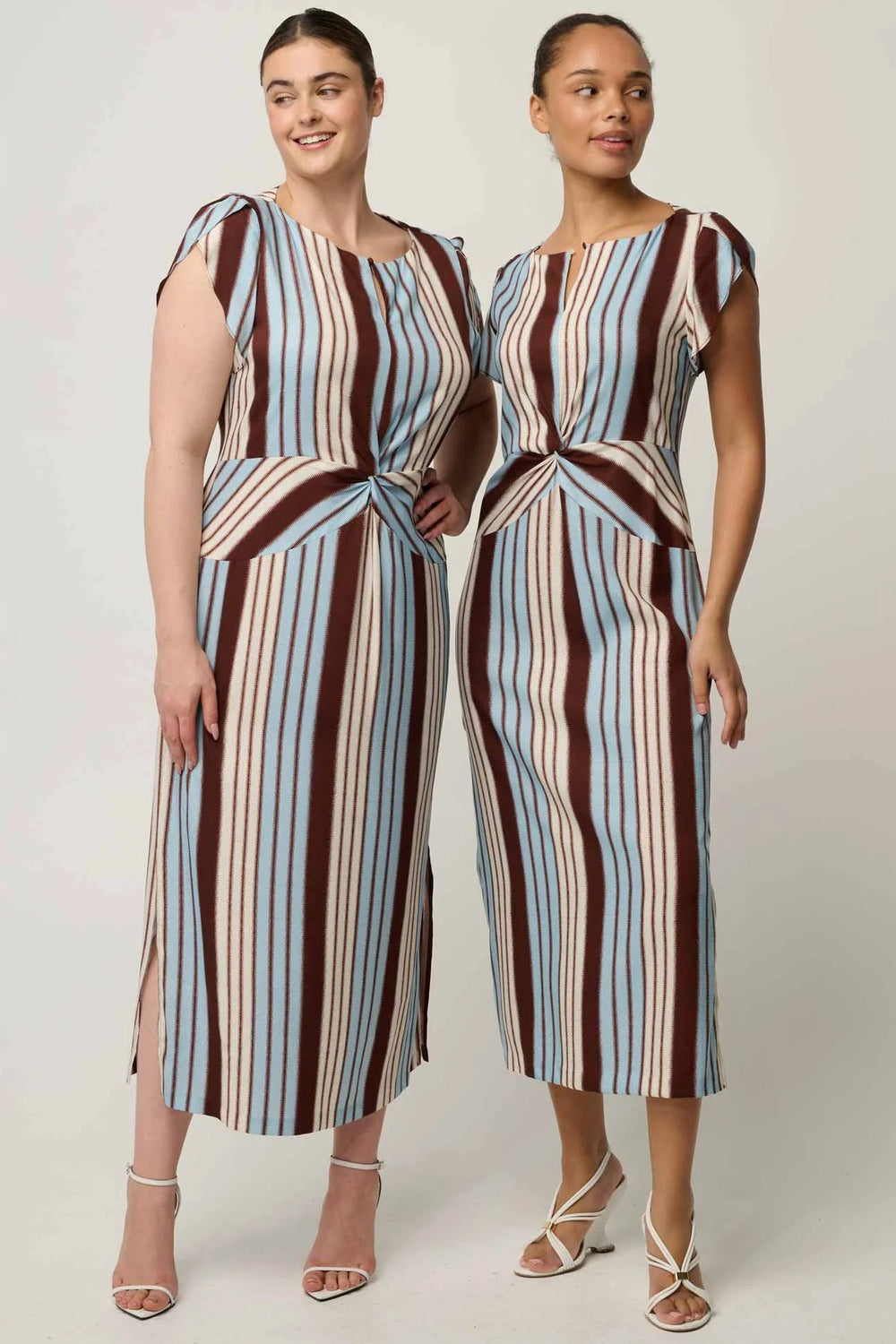 Two women modeling matching sleeveless midi dresses with vertical stripes in brown, light blue, beige, and white, standing against a plain light gray studio background.