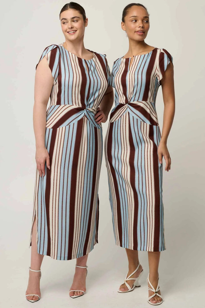 Two women modeling matching sleeveless midi dresses with vertical stripes in brown, light blue, beige, and white, standing against a plain light gray studio background.