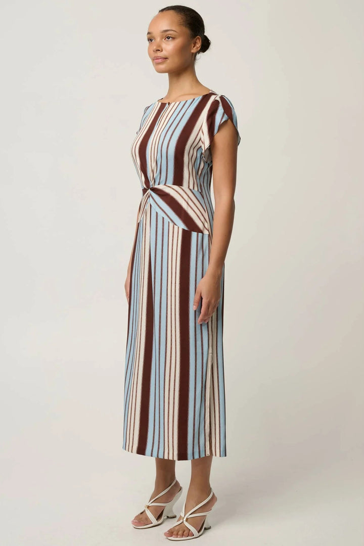 A woman modeling a midi wrap dress with vertical stripes in brown, beige, and light blue, paired with white strappy heeled sandals, against a plain light gray studio background.