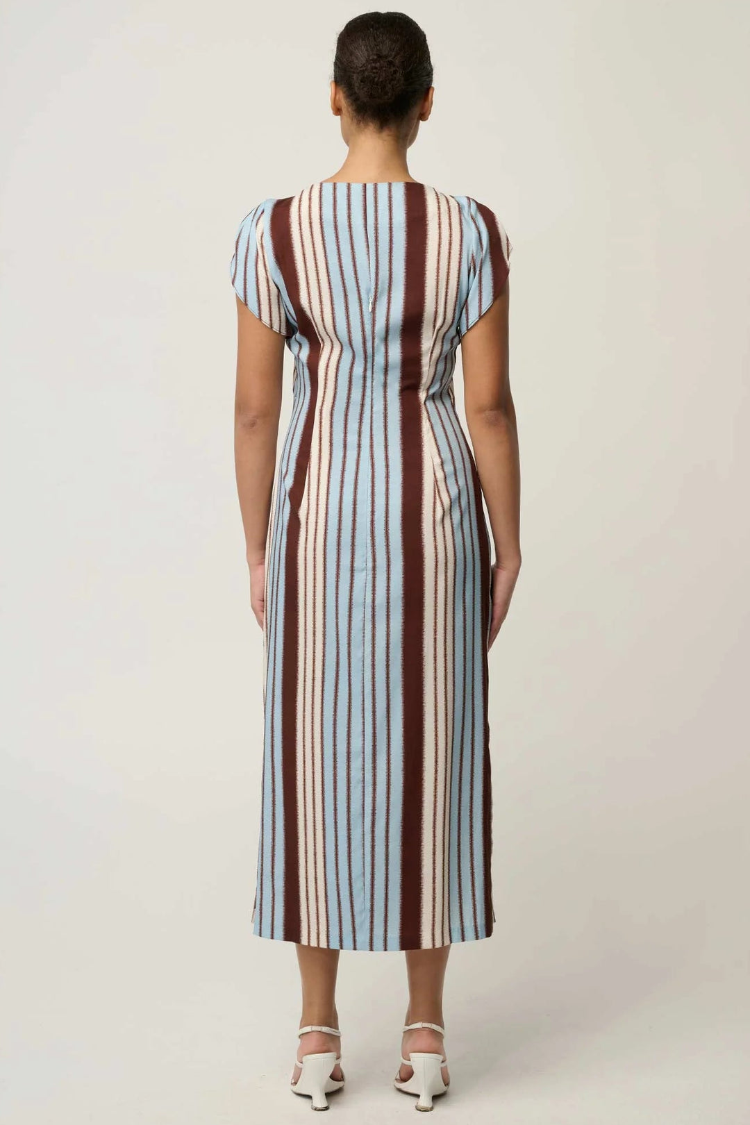 A woman viewed from the back wearing a midi dress with vertical stripes in light blue, brown, and cream, paired with white heeled sandals, against a plain light gray studio background.