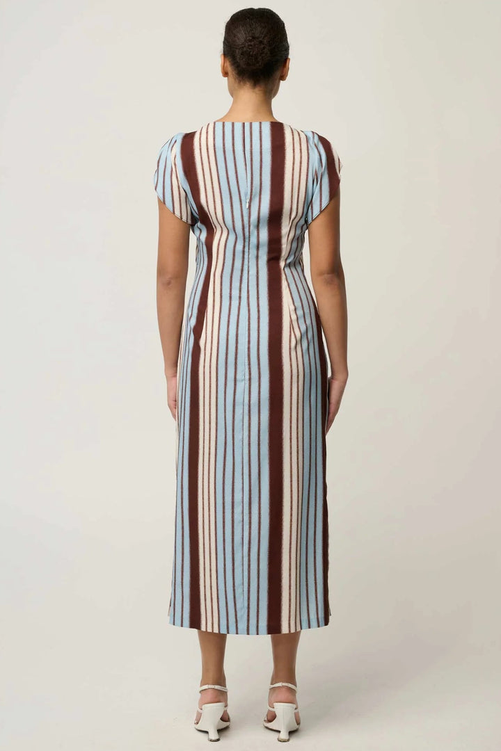 A woman viewed from the back wearing a midi dress with vertical stripes in light blue, brown, and cream, paired with white heeled sandals, against a plain light gray studio background.