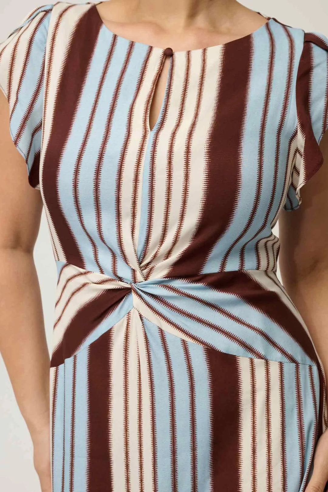 A close-up of a woman wearing a sleeveless dress with vertical brown, cream, and light blue stripes, featuring a twisted knot detail at the waist and a small keyhole cutout at the neckline.