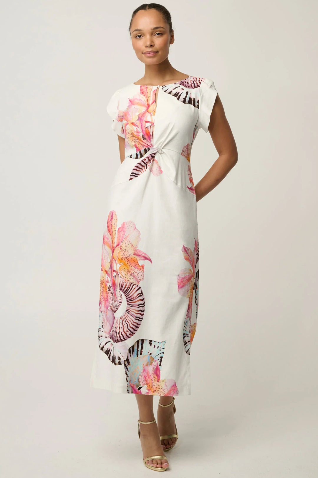 A woman modeling a white midi dress with a vibrant pink, orange, and purple floral print, styled with beige strappy high-heeled sandals against a light gray studio background.