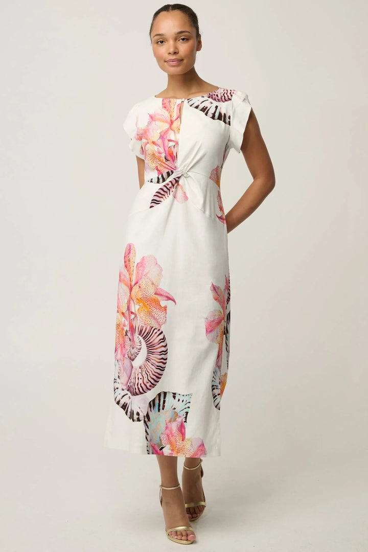 A woman modeling a white midi dress with a vibrant pink, orange, and purple floral print, styled with beige strappy high-heeled sandals against a light gray studio background.