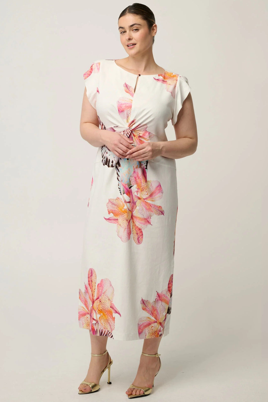 A woman modeling a white midi dress with pink and orange floral prints, featuring a twisted front detail and cap sleeves, paired with gold high-heeled sandals against a light gray studio background.