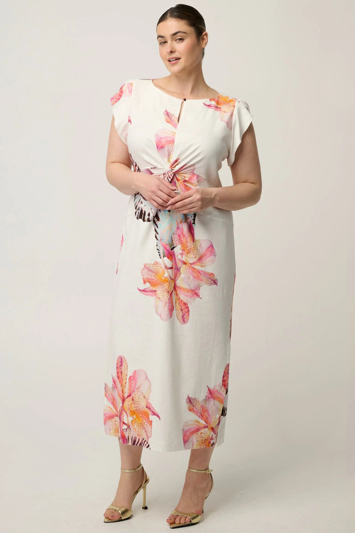 A woman modeling a white midi dress with pink and orange floral prints, featuring a twisted front detail and cap sleeves, paired with gold high-heeled sandals against a light gray studio background.
