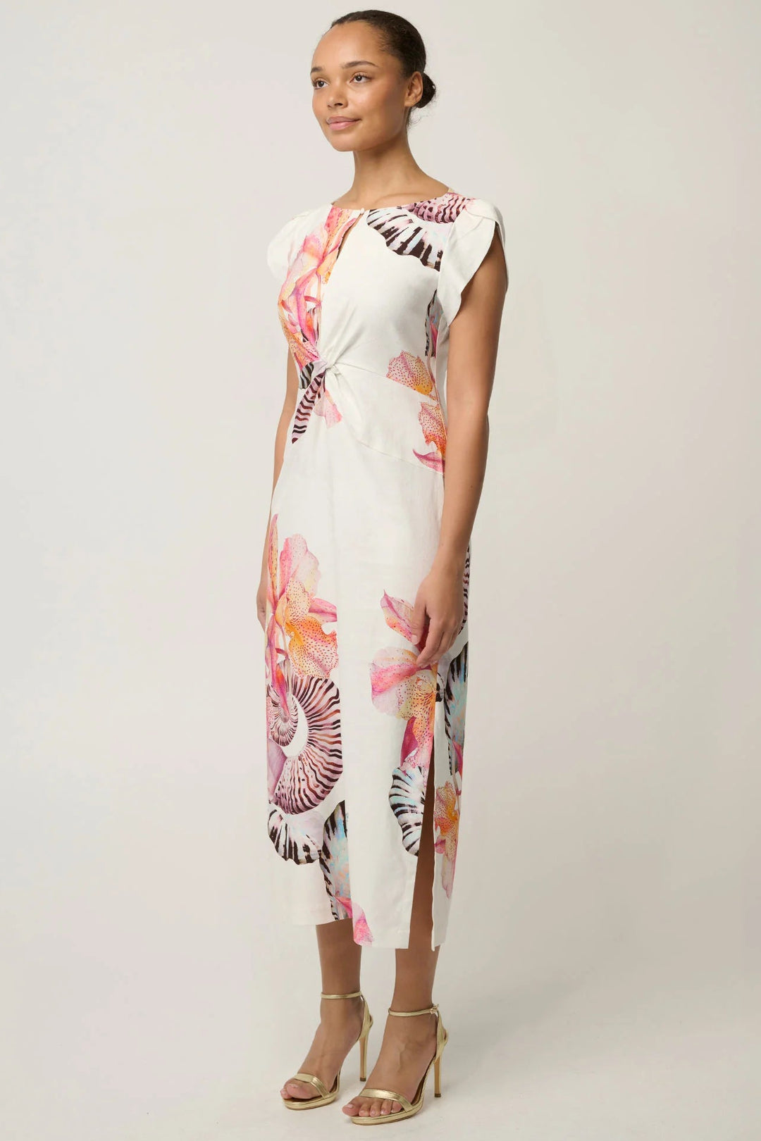 A woman modeling a white midi dress with a vibrant pink, orange, and purple floral print, paired with gold high-heeled sandals, set against a plain light gray studio background.