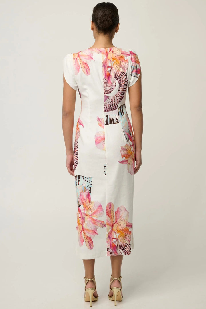 A woman viewed from the back wearing a white knee-length dress with a pink, orange, and black floral print. She is wearing beige high-heeled sandals against a plain light gray studio background.