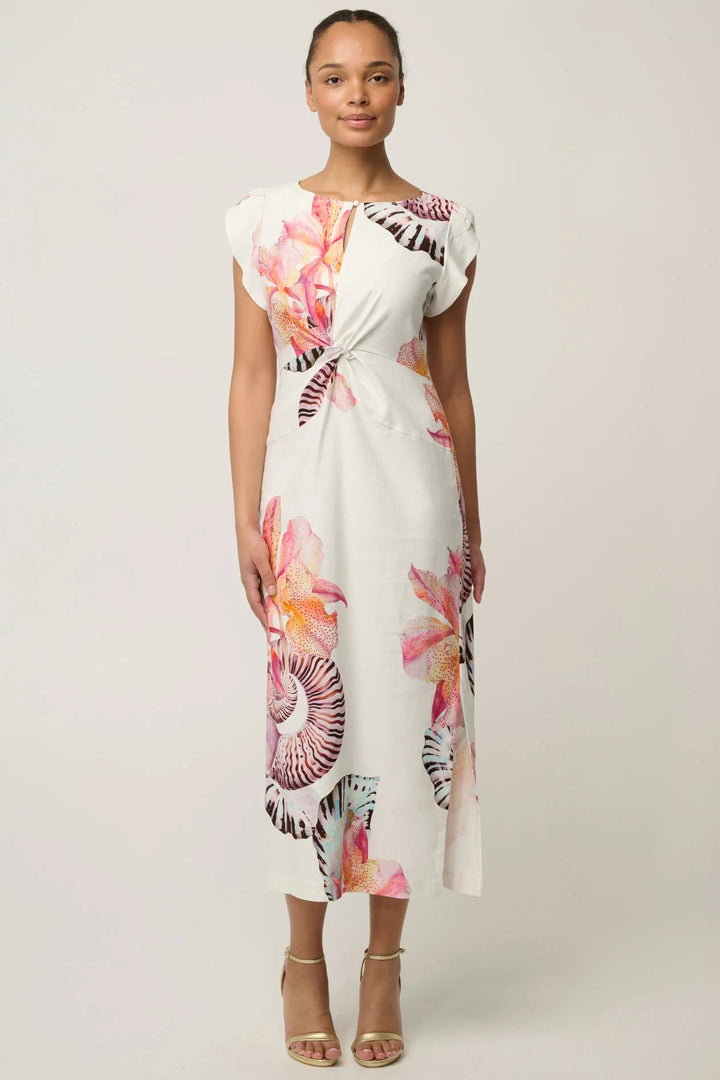 A woman modeling a white midi dress with a vibrant tropical floral print in pink, orange, and purple, styled with gold high-heeled sandals against a light gray studio background.