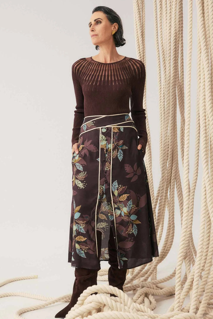 Woman wearing a brown top and patterned skirt standing next to large ropes.