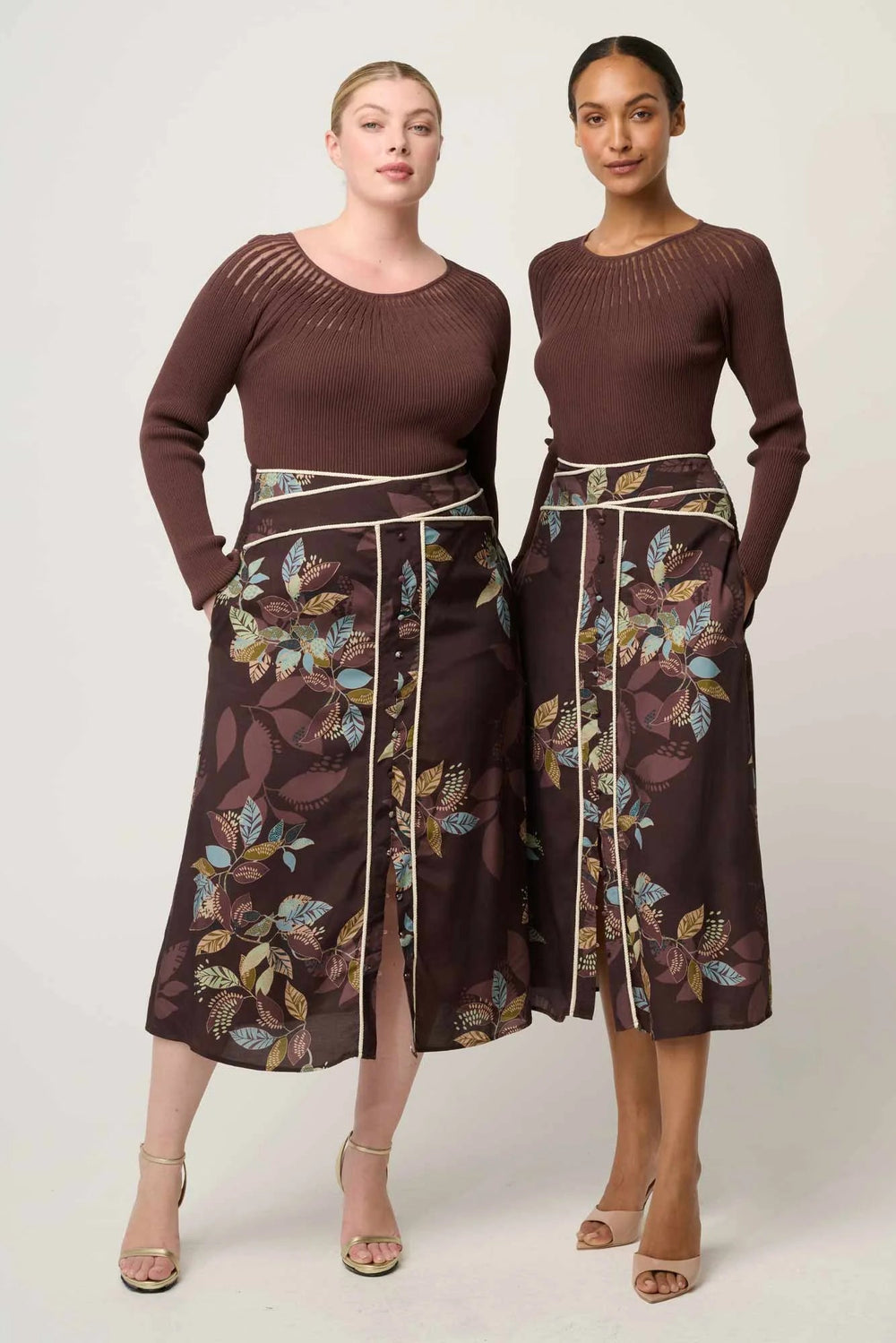 Two women wearing matching brown tops and floral skirts on a white background