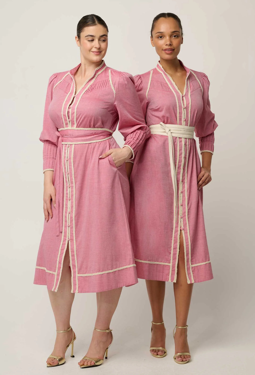 Two women modeling matching pink long-sleeve kaftan-style dresses with cream trim and white belts against a plain light gray studio background.