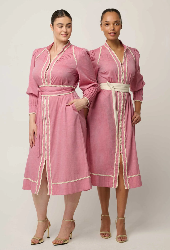 Two women modeling matching pink long-sleeve kaftan-style dresses with cream trim and white belts against a plain light gray studio background.