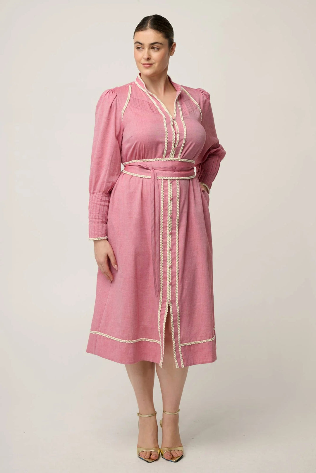A woman modeling a pink long-sleeve midi dress with cream-colored trim and a matching tie belt, set against a plain light beige studio background.