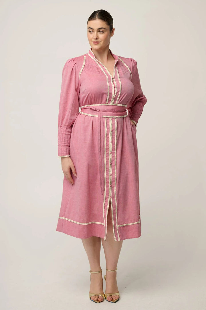 A woman modeling a pink long-sleeve midi dress with cream-colored trim and a matching tie belt, set against a plain light beige studio background.
