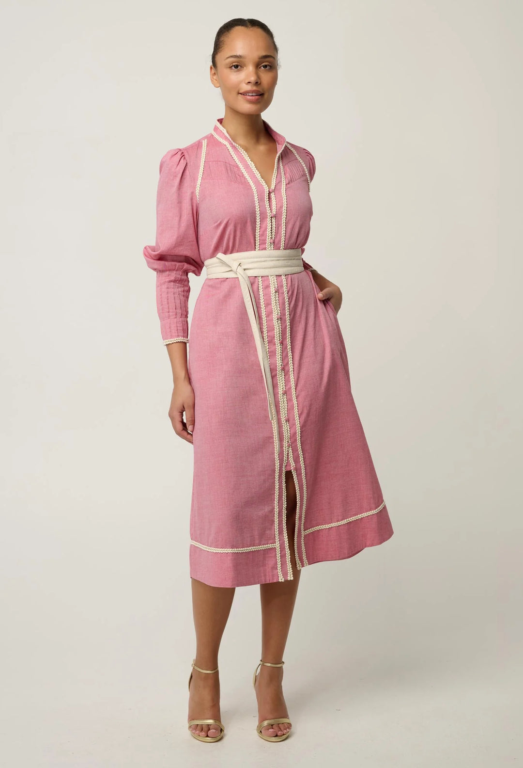 A woman modeling a pink midi shirt dress with cream trim and a matching cream belt, standing against a light gray studio background.