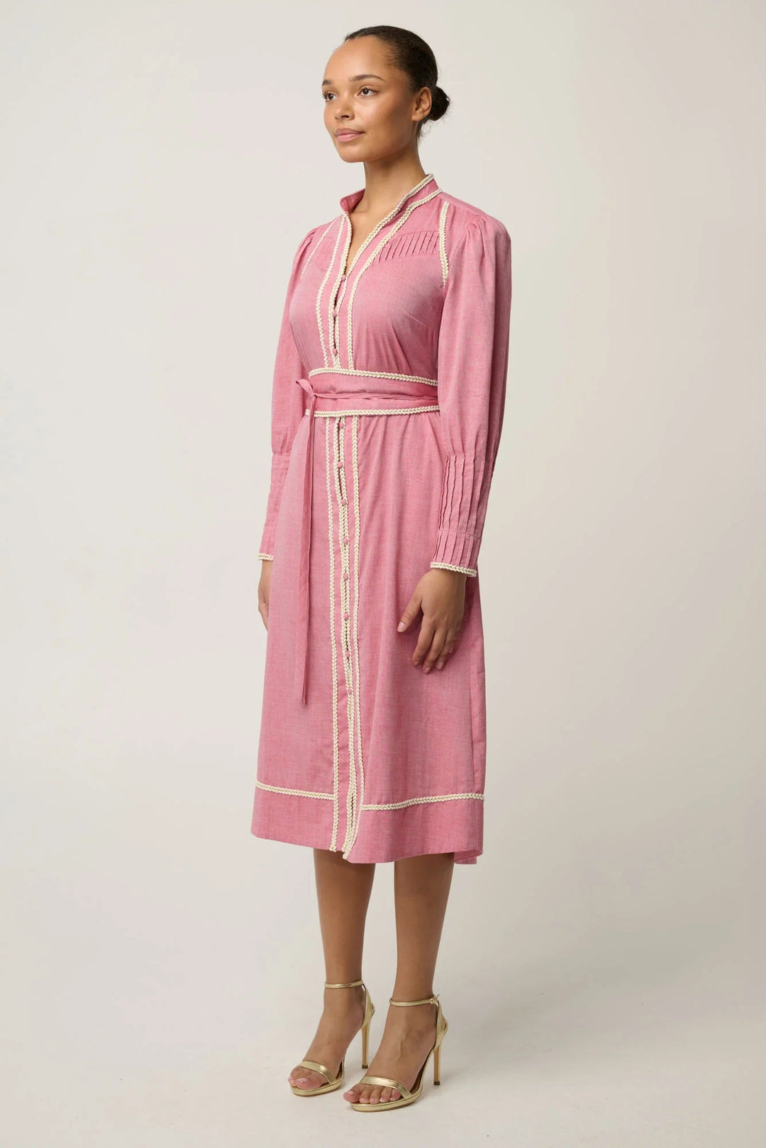 A woman modeling a pink long-sleeve midi wrap dress with white contrast piping and a matching waist tie, paired with beige high-heeled sandals against a light gray studio background.