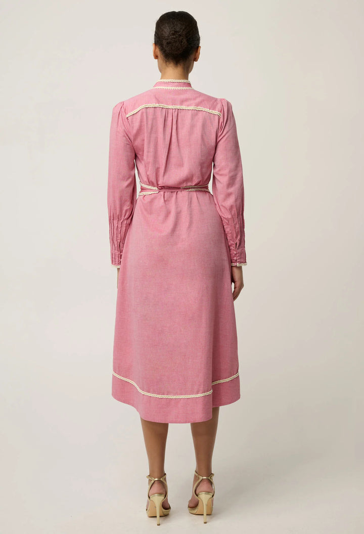 A woman viewed from the back wearing a pink long-sleeve midi dress with cream piping details, paired with beige high-heeled sandals, against a light gray studio background.