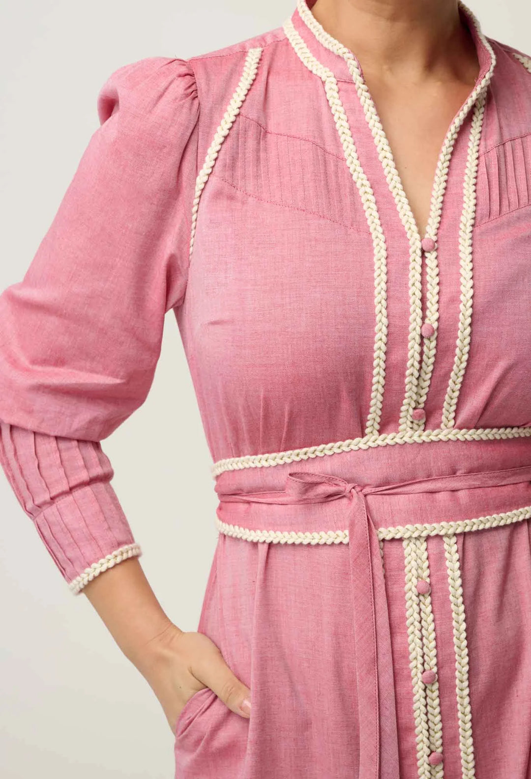 A close-up of a woman wearing a pink long-sleeve dress with cream braided trim along the neckline, sleeves, waist, and front pockets.