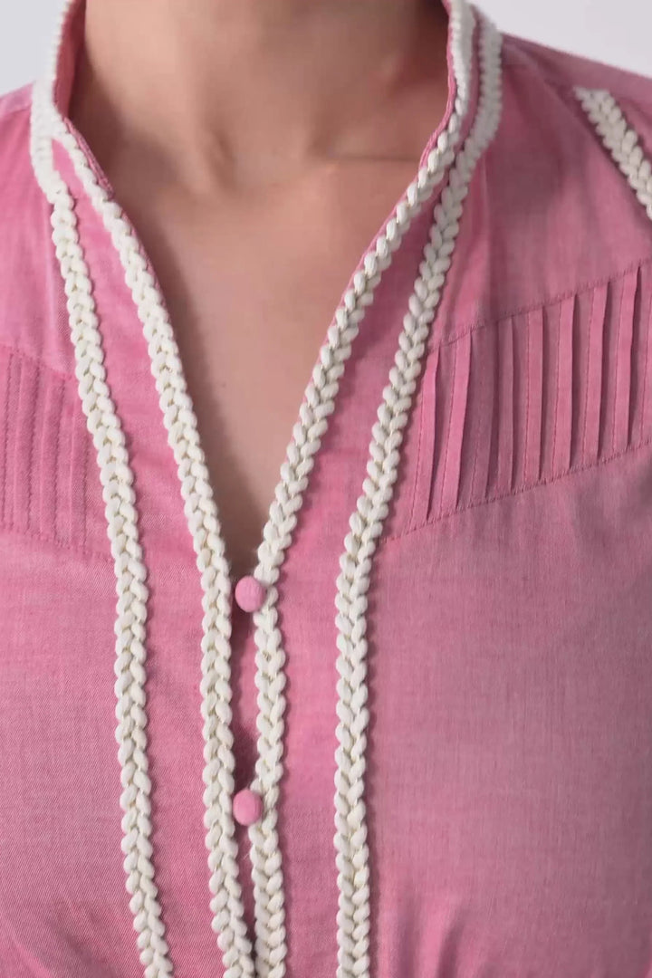 Close-up of a pink blouse with cream braided trim along the split neckline and front placket, featuring small pink buttons.