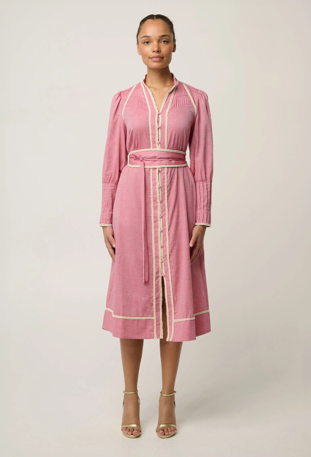 A woman modeling a pink long-sleeve midi dress with cream-colored piping and a matching tie belt, standing against a light gray studio background.