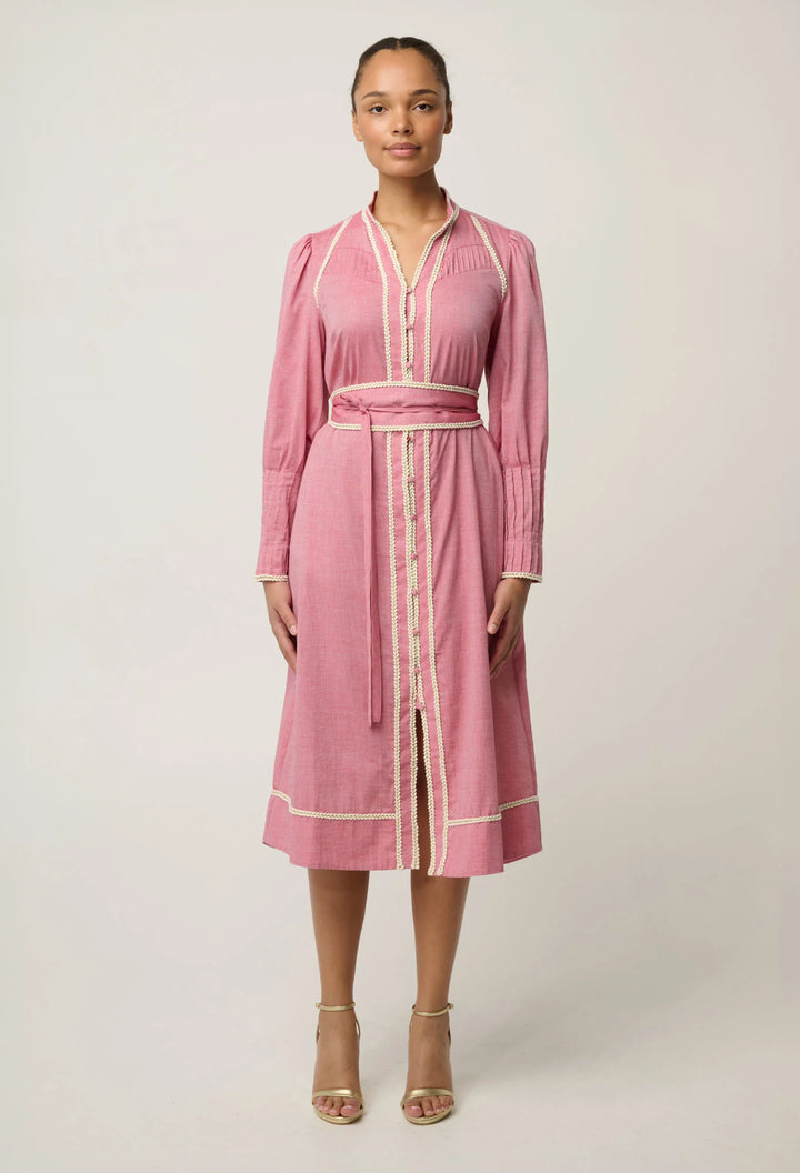 A woman modeling a pink long-sleeve midi dress with cream-colored piping and a matching tie belt, standing against a light gray studio background.