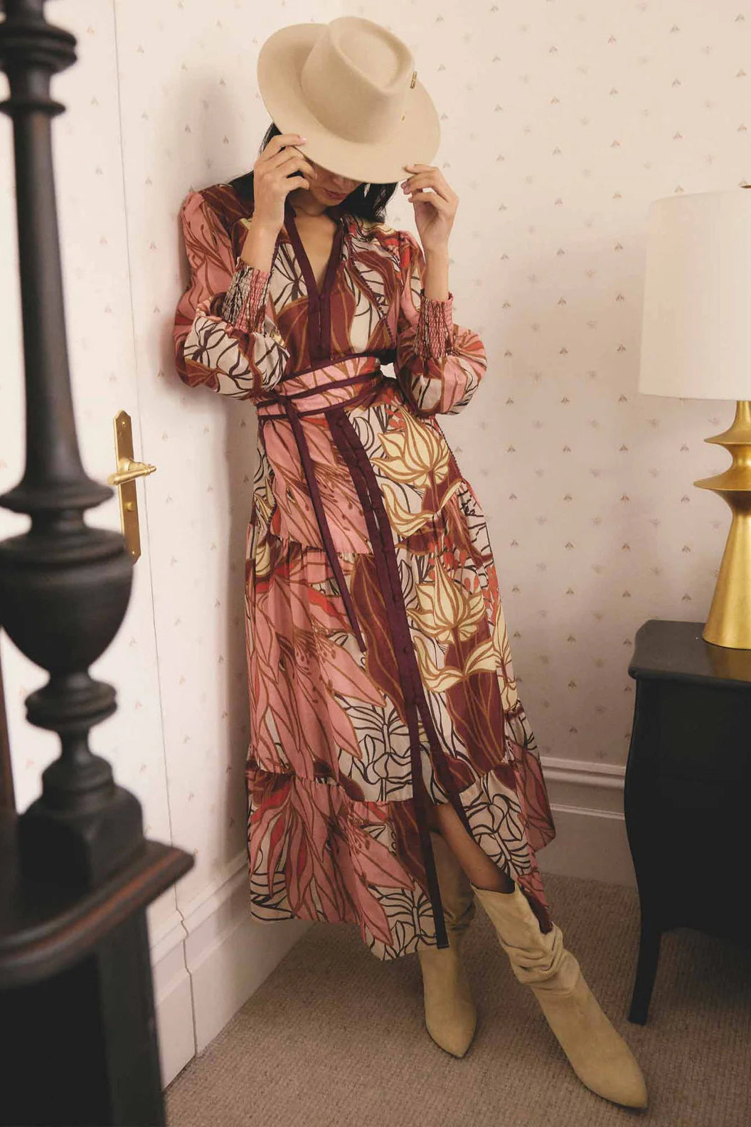 Person wearing a floral dress and beige hat in a room with a lamp and door.
