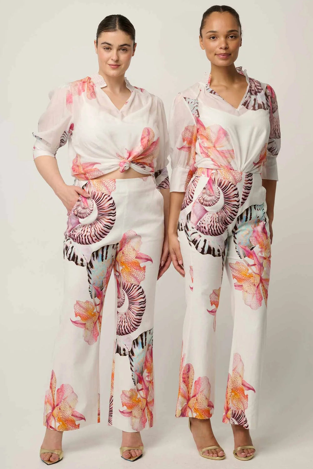 Two models wearing matching white two-piece jumpsuits with a vibrant pink, orange, and purple floral print, standing against a light gray studio background.