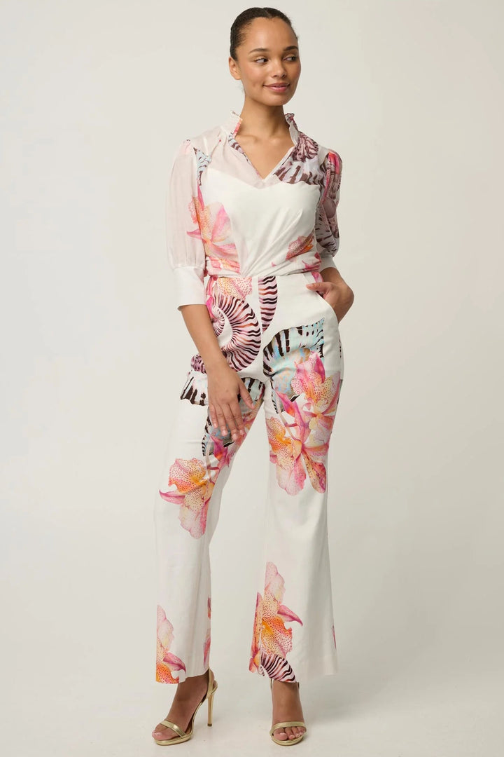 A woman modeling a white jumpsuit with a vibrant pink, orange, and black floral print, styled with clear sheer puff sleeves and matching tie-waist sash, paired with nude high-heeled sandals against a light gray studio background.