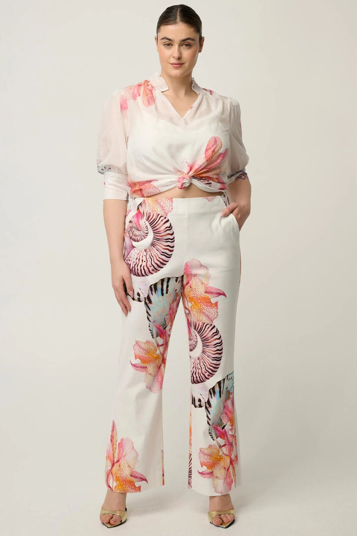 A woman modeling a matching two-piece set featuring a sheer white top with pink floral prints and white wide-leg trousers with large pink, orange, and purple floral and shell-like patterns, set against a plain light gray studio background.