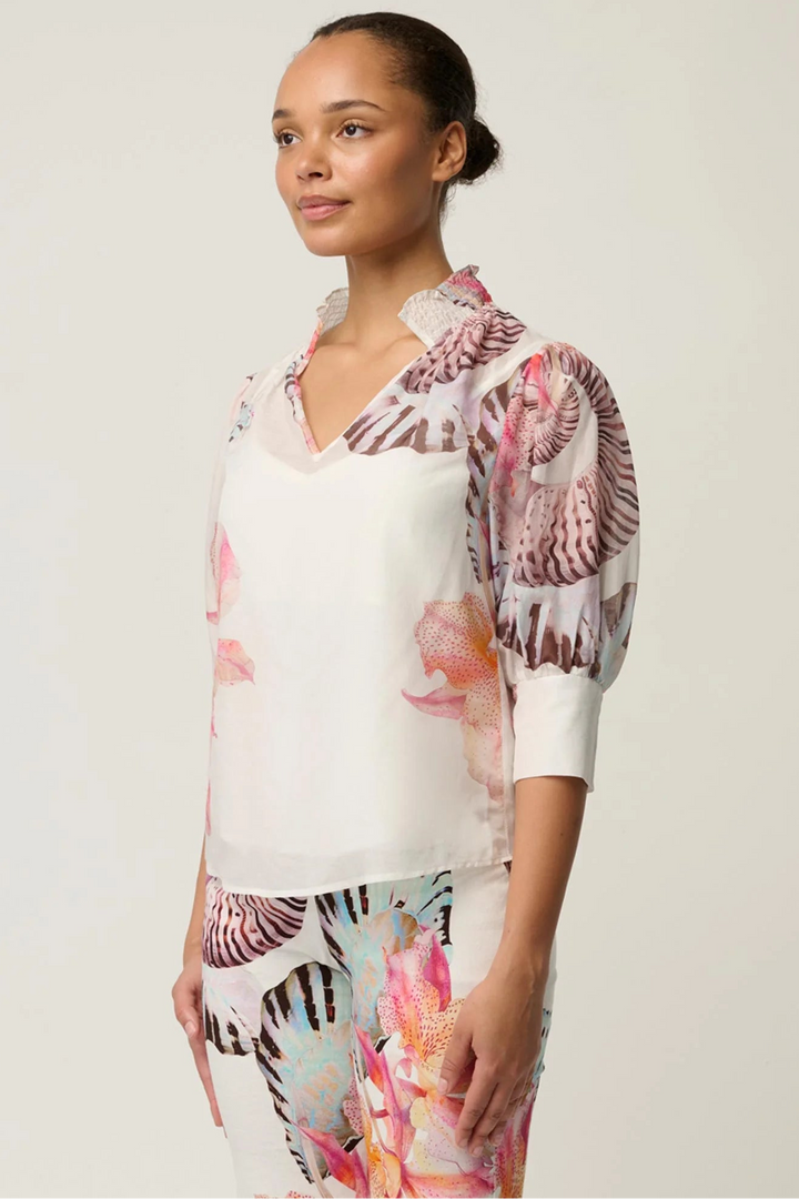 A woman modeling a white blouse and matching skirt set featuring a vibrant pink, orange, and brown tropical floral print on the sleeves and lower portions.
