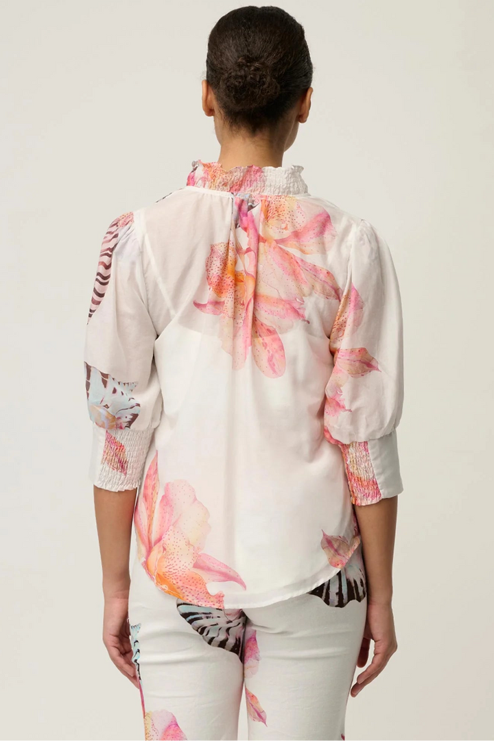 A woman viewed from the back wearing a white blouse with a pink and orange floral print and patterned sleeve inserts, paired with white pants featuring a matching floral print, against a light gray studio background.
