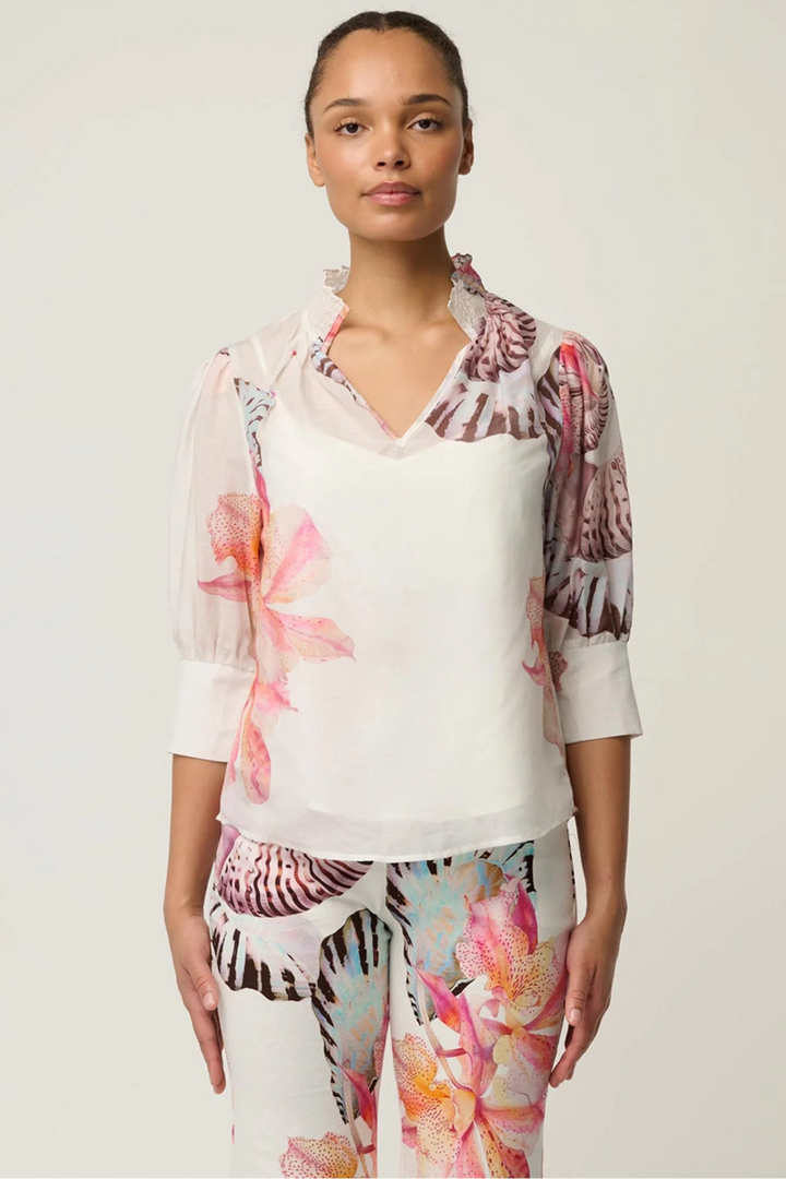 A model wearing a white blouse with pink and brown floral patterns on the sleeves and collar, paired with matching patterned trousers, against a light gray studio background.