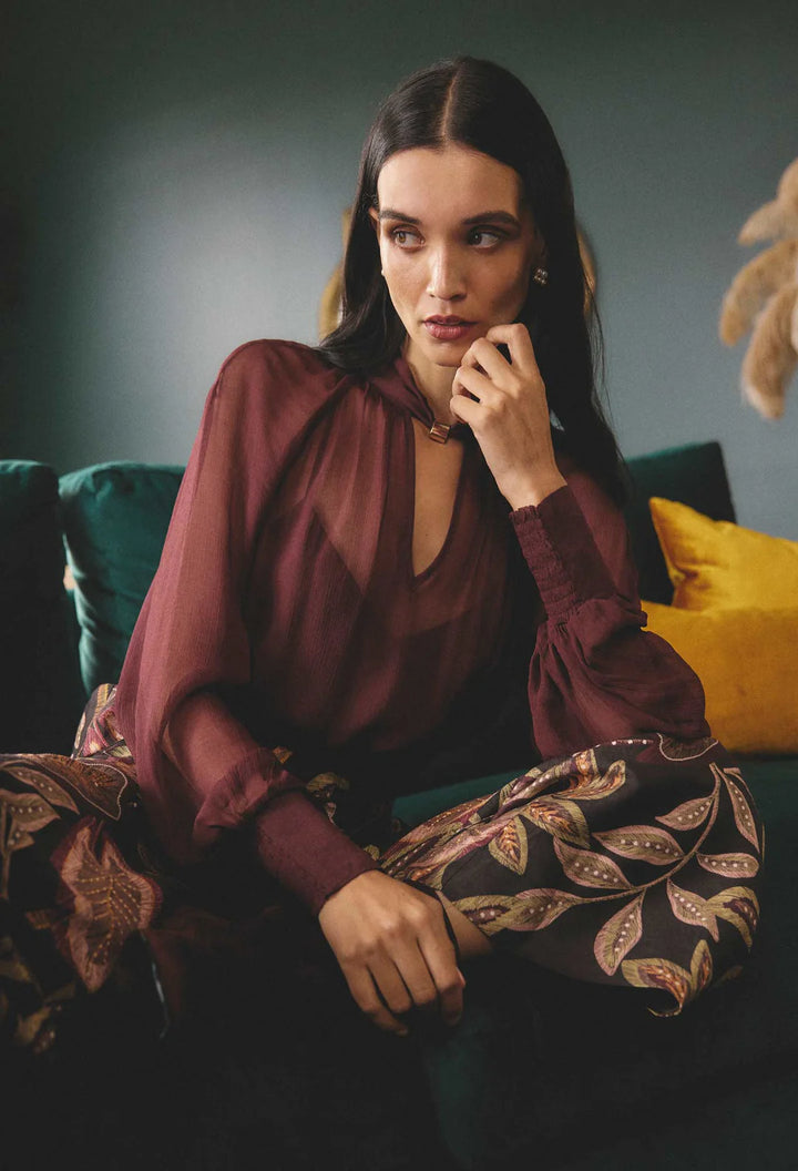 Woman sitting on a couch wearing a burgundy blouse and patterned pants.