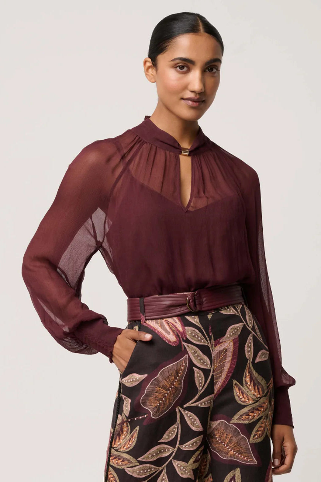 Woman wearing a burgundy blouse with sheer sleeves and patterned pants on a light gray background