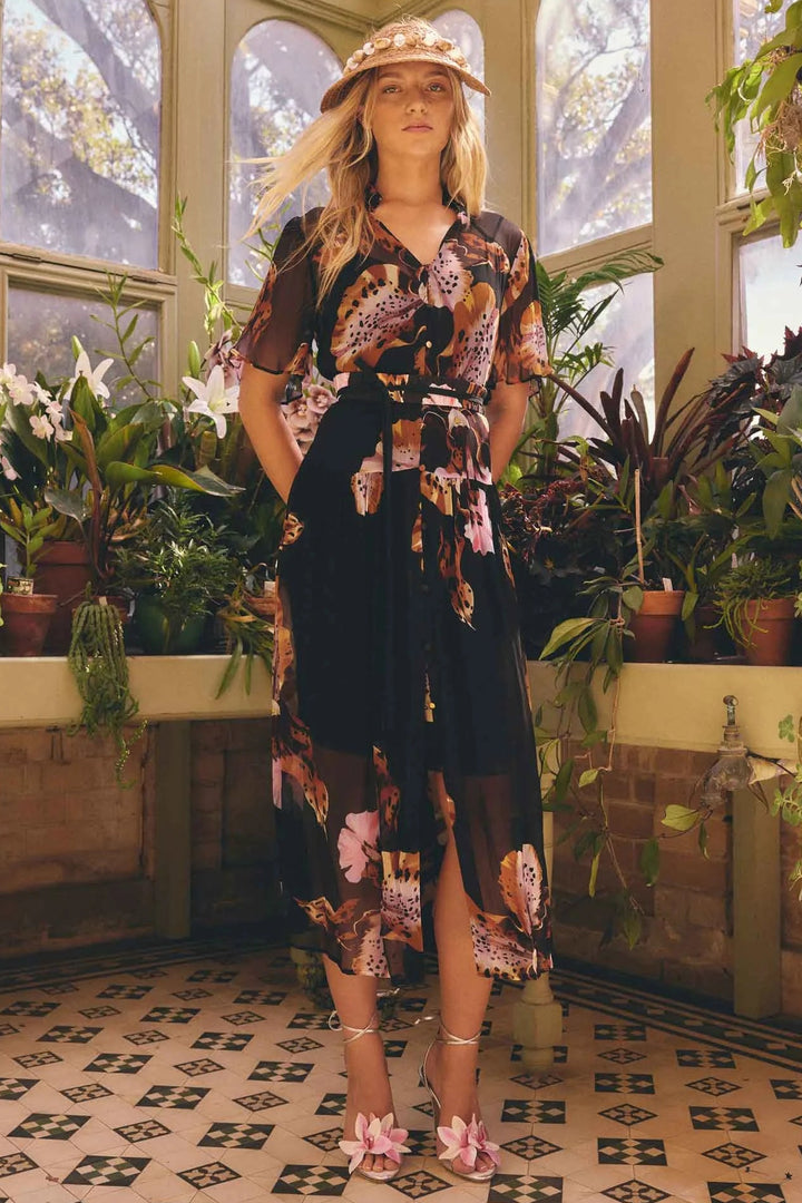 A woman stands in a sunlit greenhouse wearing a sheer black midi dress with a pink, brown, and beige floral print, a matching hat, and pink heels.