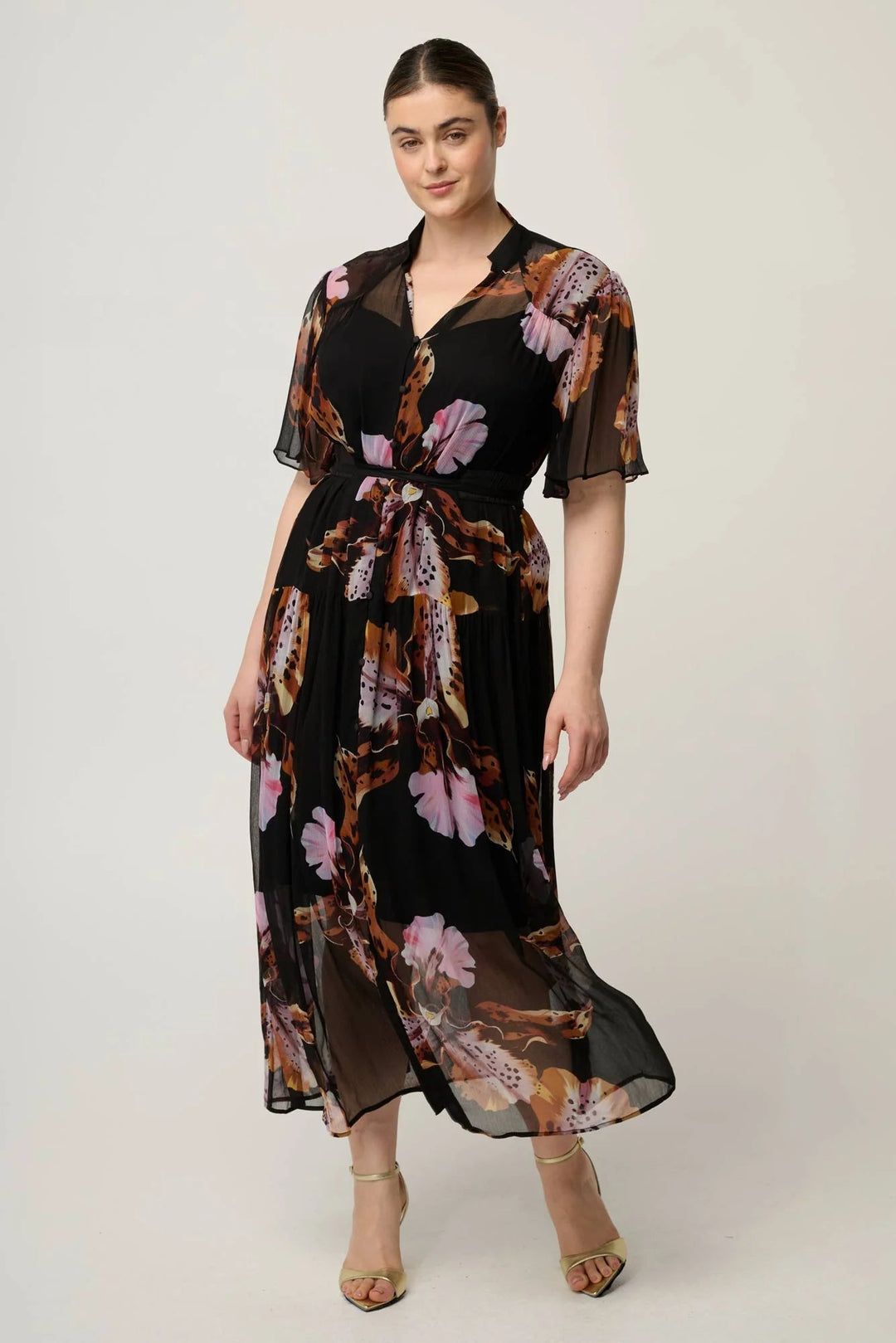 A woman modeling a black midi dress with a sheer floral print in pink, brown, and beige tones, featuring short flutter sleeves and a tie waist, set against a plain light gray studio background.