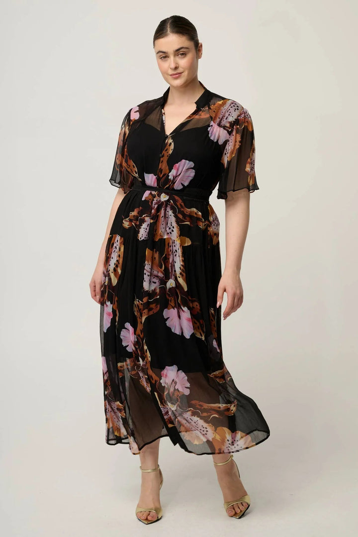 A woman modeling a black midi dress with a sheer floral print in pink, brown, and beige tones, featuring short flutter sleeves and a tie waist, set against a plain light gray studio background.
