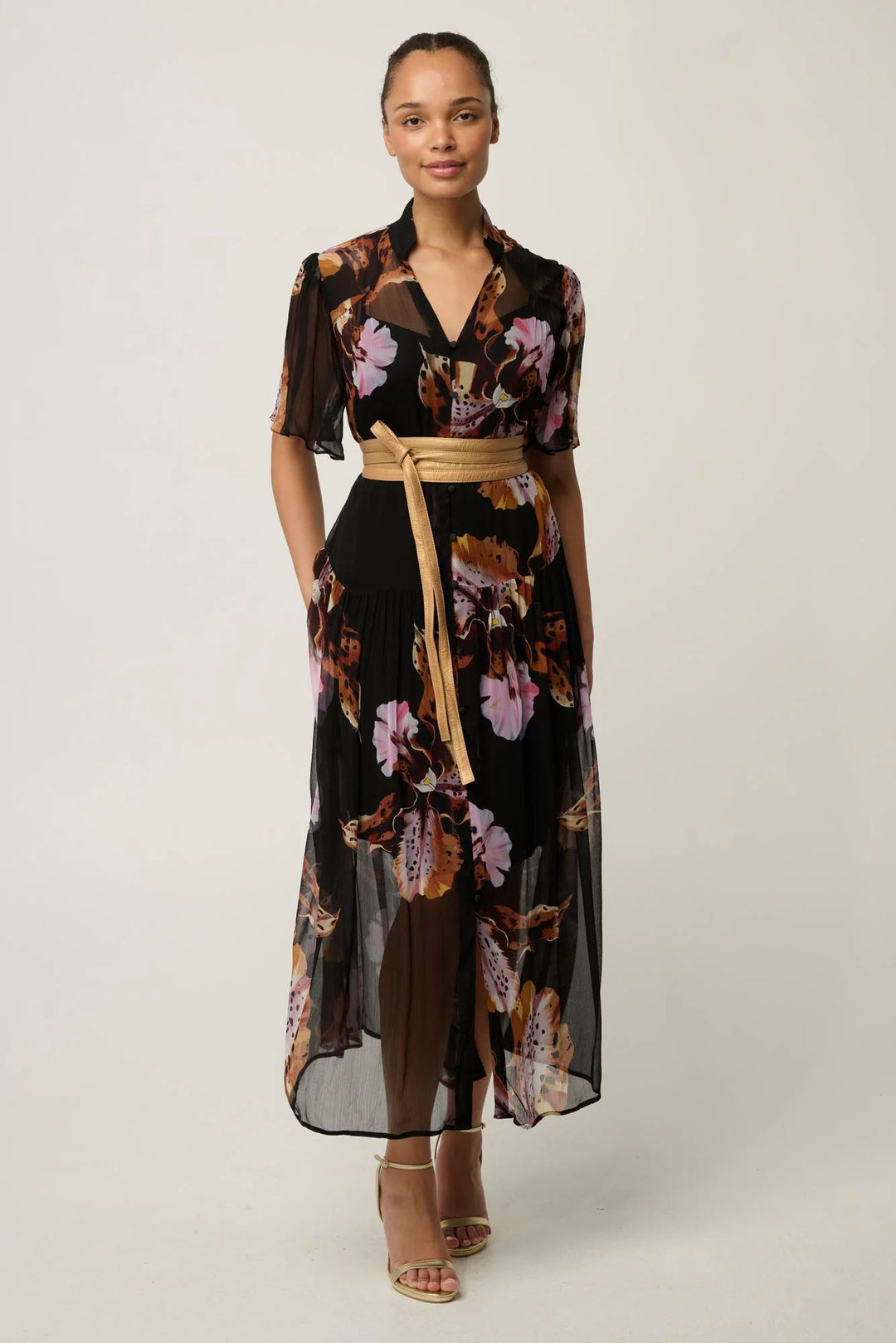 A woman modeling a black midi wrap dress with a brown, pink, and purple floral print, featuring short sleeves, a V-neckline, and a tan belt.