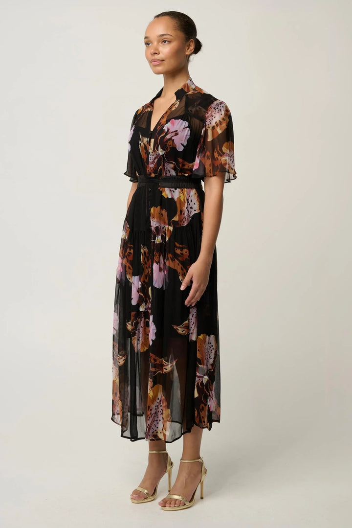 A woman modeling a black midi dress with a brown, pink, and purple floral print, featuring short flutter sleeves and a tie waist, set against a plain light gray studio background.