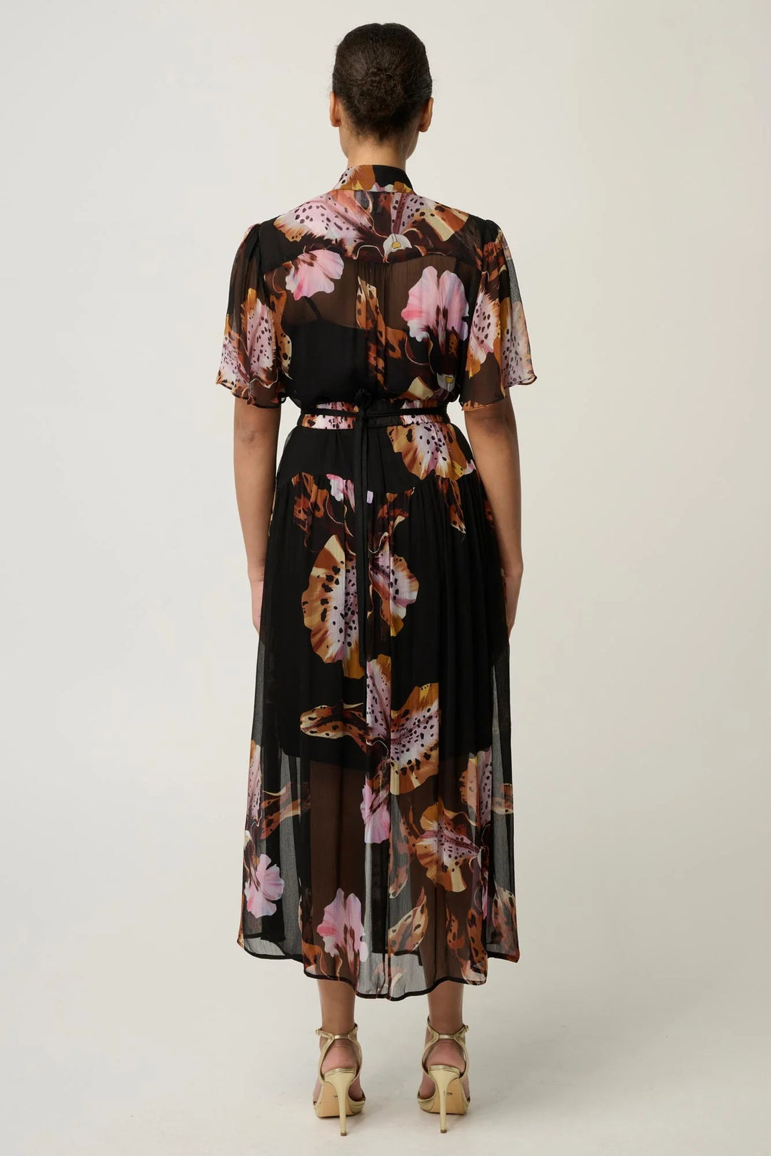 A woman viewed from the back wearing a black midi dress with a pink, brown, and beige floral print, paired with beige high-heeled sandals, against a light gray studio background.