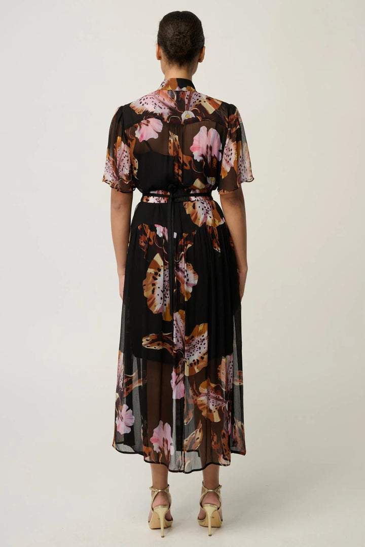 A woman viewed from the back wearing a black midi dress with a pink, brown, and beige floral print, paired with beige high-heeled sandals, against a light gray studio background.