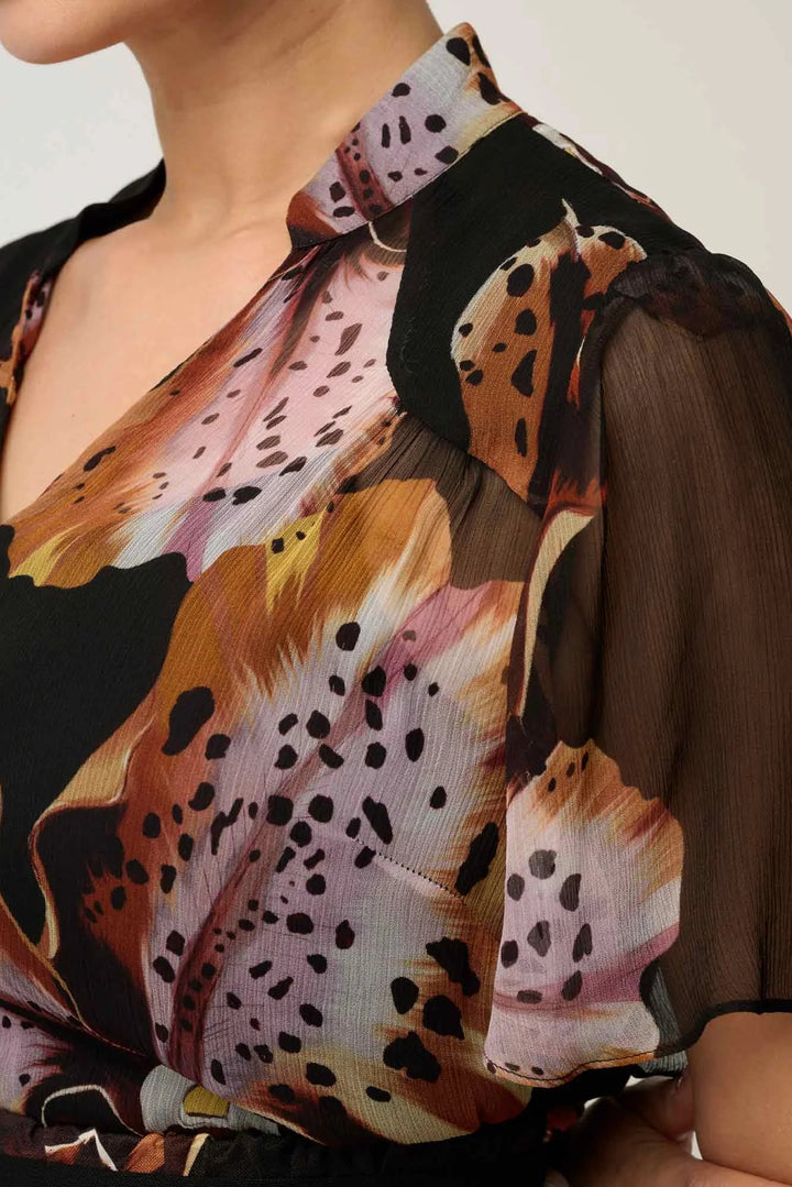 A close-up of a woman wearing a sheer, semi-sheer blouse with a bold abstract print in brown, orange, pink, black, and white. The blouse features a V-neckline and short sleeves with a sheer overlay.