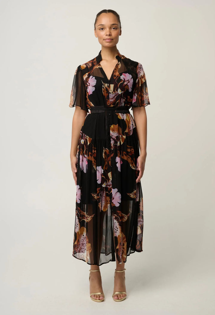 A model stands against a light gray studio background wearing a black midi dress with a brown, pink, and purple floral print, sheer flutter sleeves, and a matching black belt.