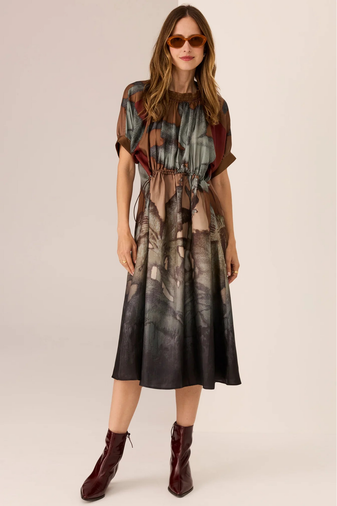 Woman wearing a patterned dress with sheer sleeves and a belt, standing against a plain background.